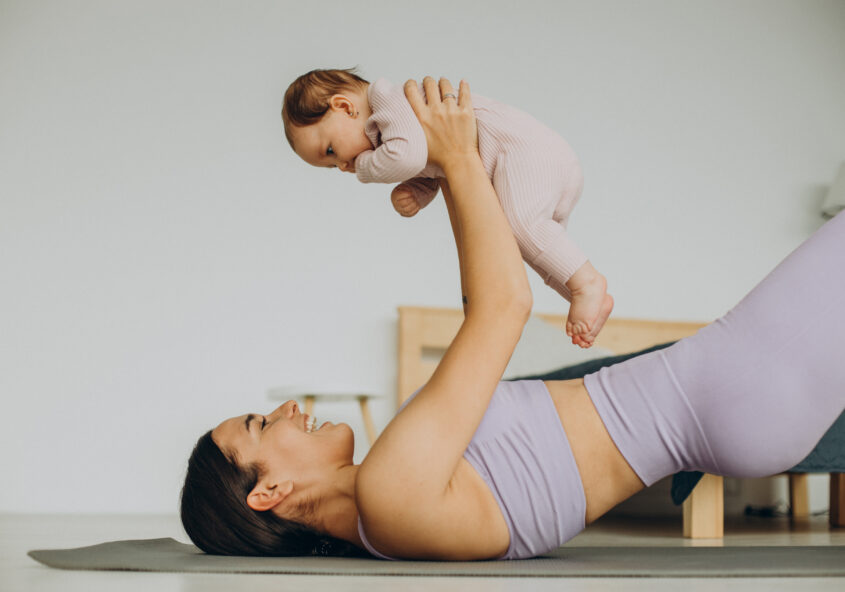 Parent lying on back on a mat, lifting a smiling baby wearing a pink onesie above them in a playful moment at home