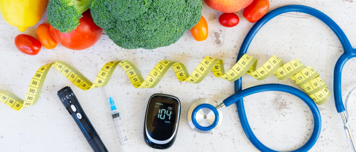 Healthy eating and health monitoring scene with fruits, vegetables, a measuring tape, and medical devices like a stethoscope and glucose meter on a white surface.